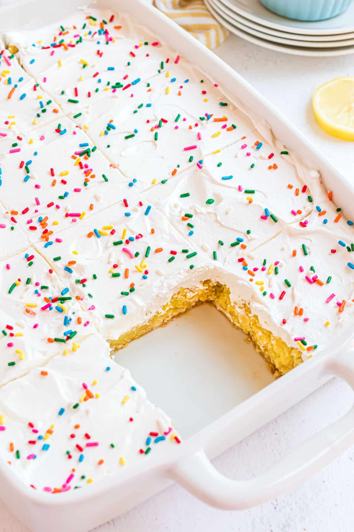 Lemon cake in a white 13x9 topped with whipped cream and sprinkles with one slice removed from pan.