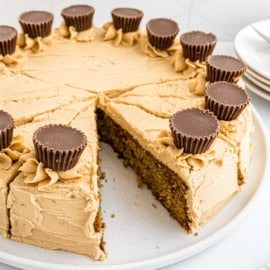 Round Reese's peanut butter cake with creamy peanut butter frosting, topped with mini peanut butter cups; one slice is missing.