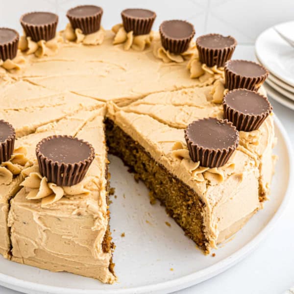 Round Reese's peanut butter cake with creamy peanut butter frosting, topped with mini peanut butter cups; one slice is missing.