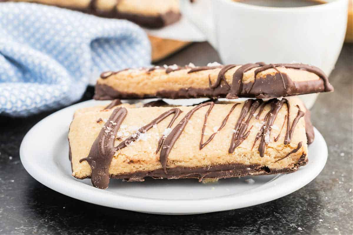 Two dark chocolate peanut butter shortbread cookies, drizzled and dipped, rest on a white plate next to a steaming cup of coffee.