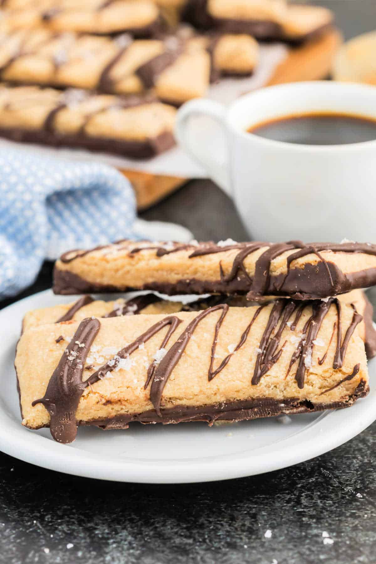 Two dark chocolate peanut butter shortbread cookies are drizzled with chocolate and served on a plate next to a steaming cup of coffee.