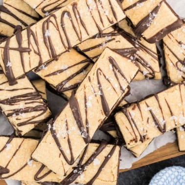 Dark chocolate peanut butter shortbread cookies drizzled with chocolate and sprinkled with sea salt, stacked on parchment paper.