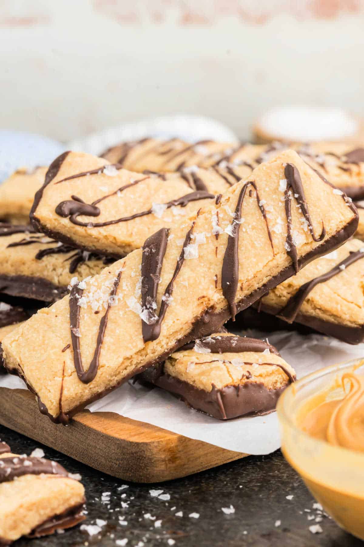 Rectangular dark chocolate peanut butter shortbread cookies drizzled with chocolate and sprinkled with sea salt, stacked on a wooden board.