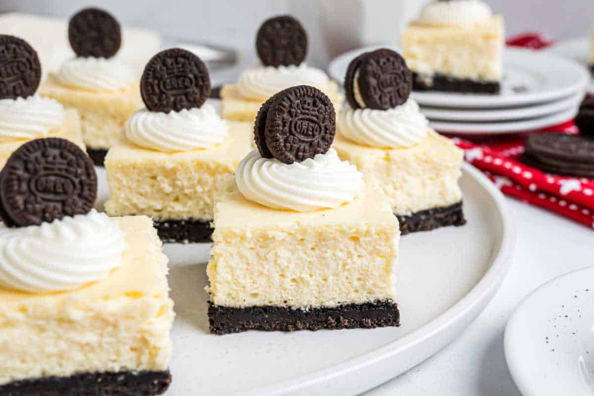 Mini cheesecake bars with an Oreo cookie crust, topped with whipped cream and a mini Oreo, arranged on a white plate—these irresistible Oreo cheesecake bites are perfect for any dessert lover.