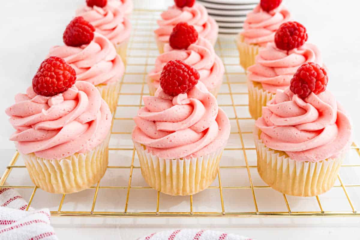Raspberry lemonade cupcakes with vanilla bases, topped with pink frosting and fresh raspberries, arranged on a wire cooling rack.