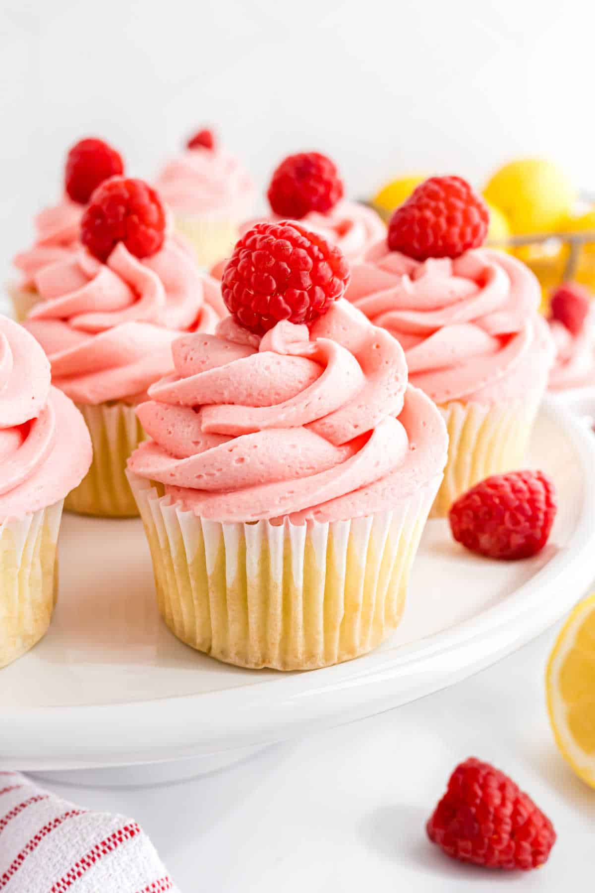 Raspberry lemonade cupcakes with vanilla bases and pink raspberry frosting, each topped with a fresh raspberry, on a white cake stand.