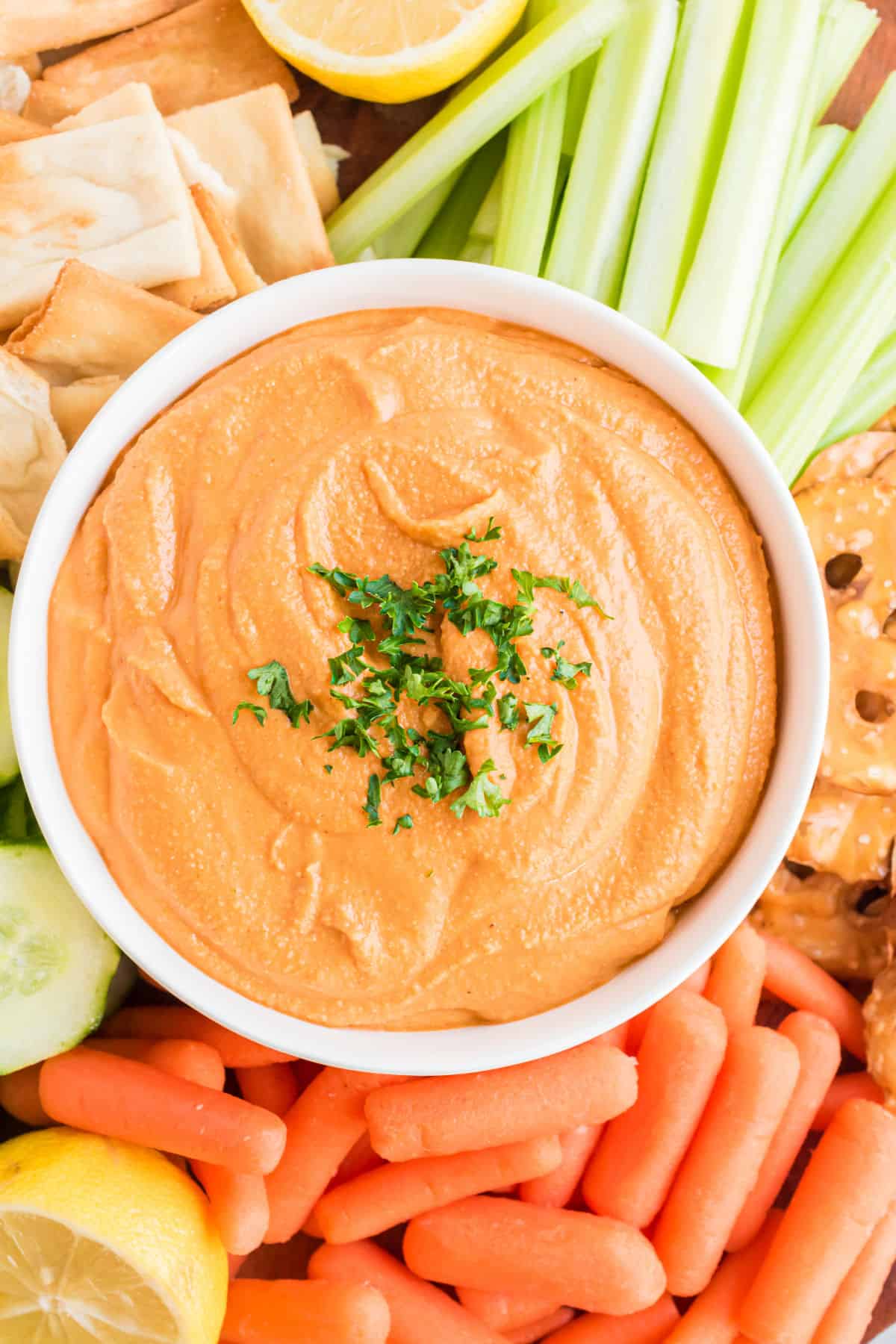 Red pepper hummus in a white bowl serve with veggies and pita chips.