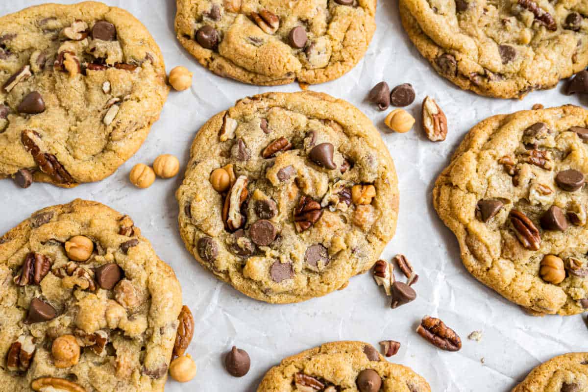 Caramel pecan chocolate chip cookies with hazelnuts rest on parchment paper, surrounded by scattered chocolate chips and nuts.