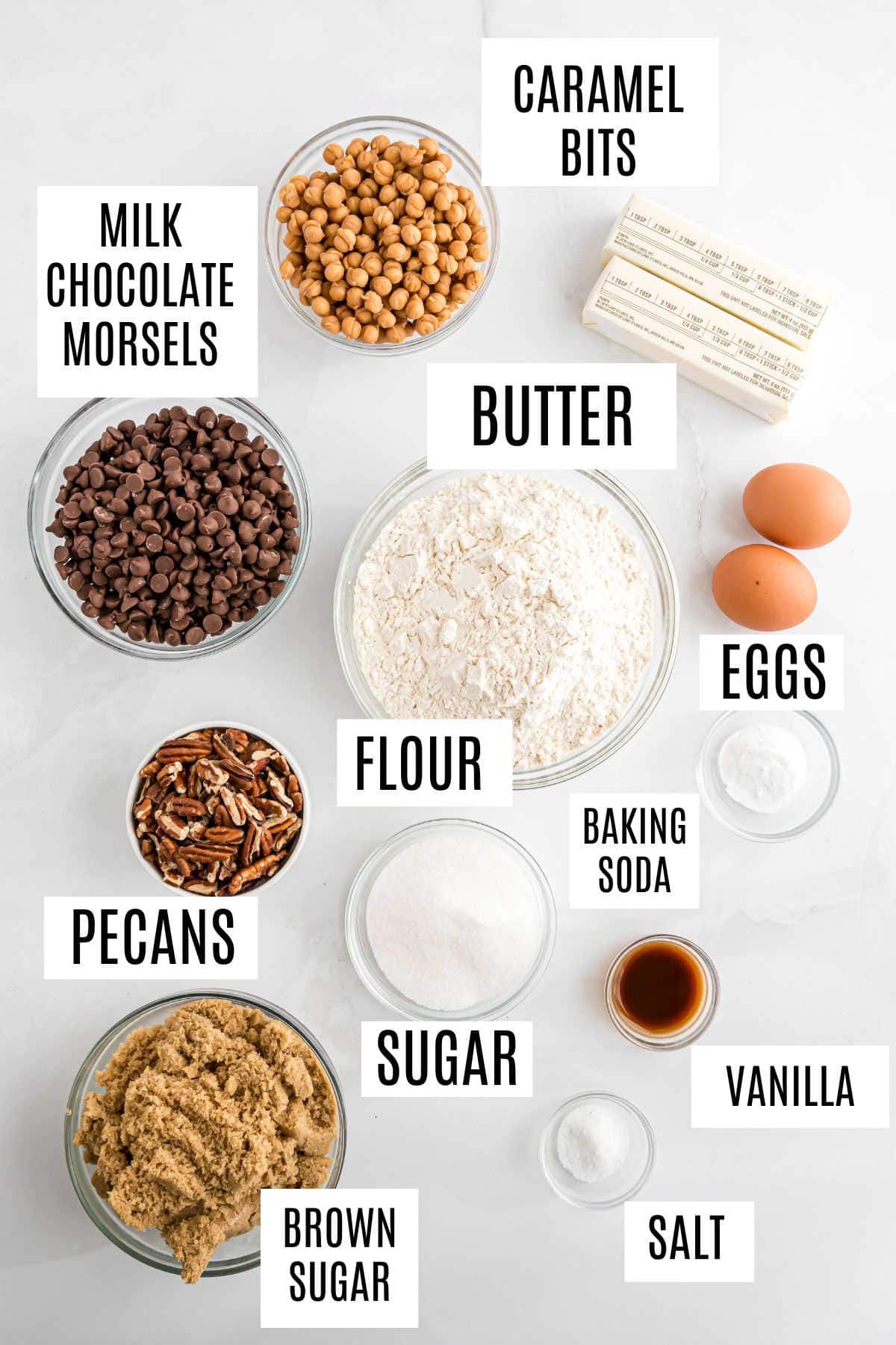 Baking ingredients on a white surface, including labeled bowls of flour, sugar, butter, eggs, chocolate, caramel, and pecans—everything you need to make delicious caramel pecan chocolate chip cookies.