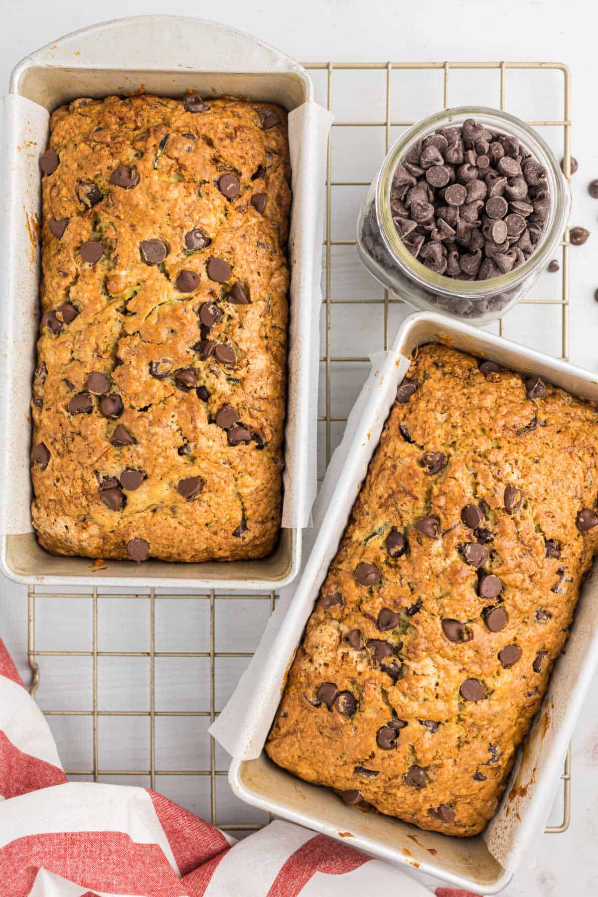 Two loaves of chocolate chip zucchini bread cool in pans on a wire rack, with a jar of chocolate chips beside them.