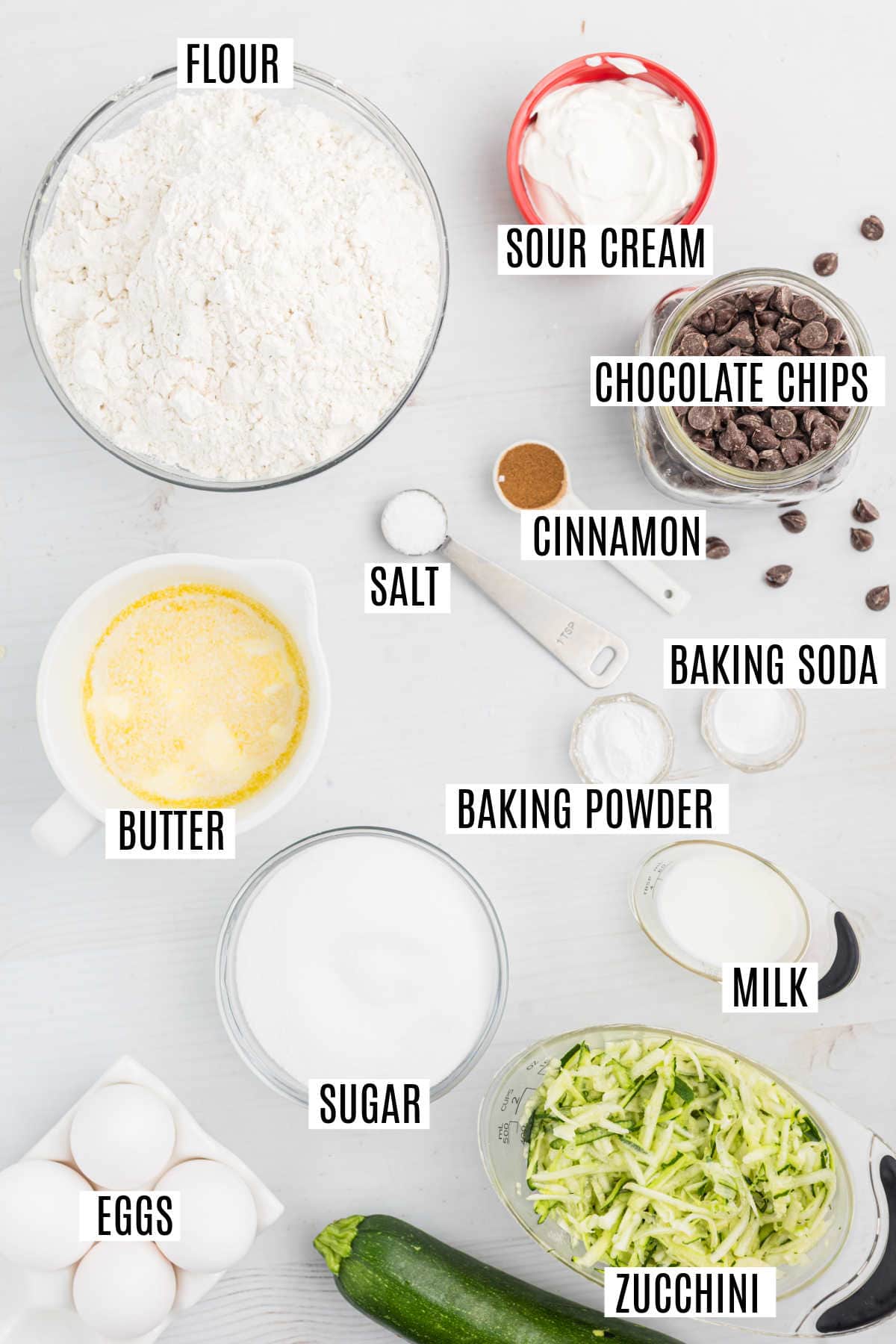 Labeled baking ingredients for chocolate chip zucchini bread—including flour, eggs, sugar, zucchini, milk, chips, butter, and spices—are arranged on a white table.