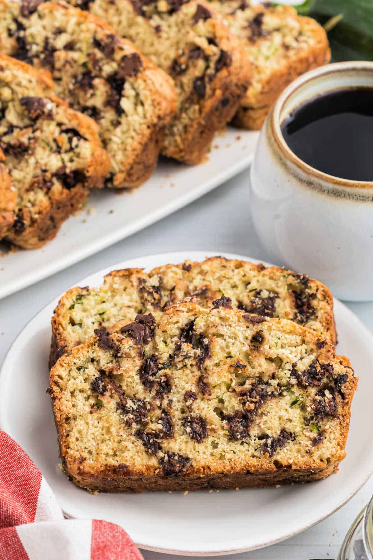 Two slices of moist chocolate chip zucchini bread sit on a plate, paired with a steaming cup of coffee and extra chocolate chip zucchini bread in the background.