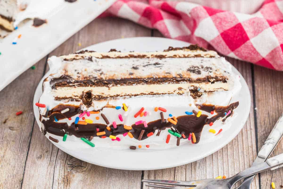 A slice of ice cream cake with chocolate sauce and rainbow sprinkles sits on a white plate, next to a fork. Indulge in this delightful ice cream cake treat for any celebration.