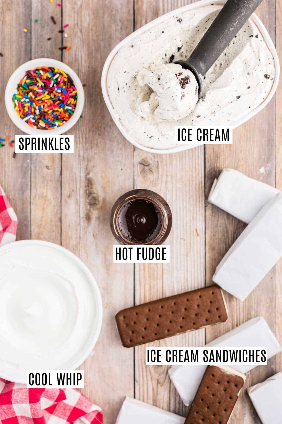 Ice cream, sprinkles, hot fudge, cool whip, and ice cream sandwiches are arranged on a wooden surface, perfect for building your own ice cream cake.
