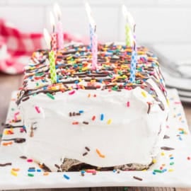 Rectangular ice cream cake with white frosting, colorful sprinkles, chocolate drizzle, and five lit candles on top.