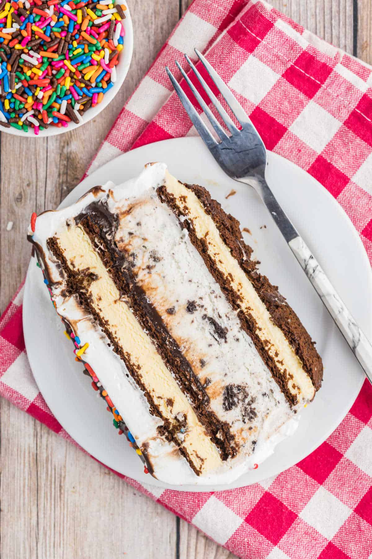 A slice of layered ice cream cake rests on a white plate, with a fork and colorful sprinkles nearby.