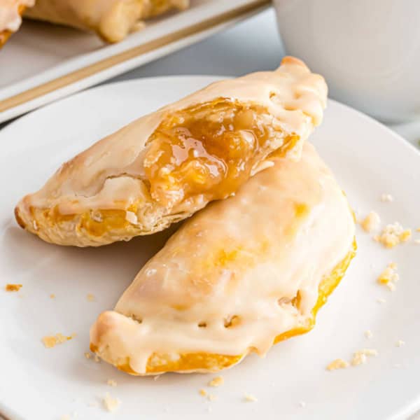 Two glazed peach hand pies on a white plate, reminiscent of classic apple empanadas, with one broken open to reveal the luscious peach filling inside.