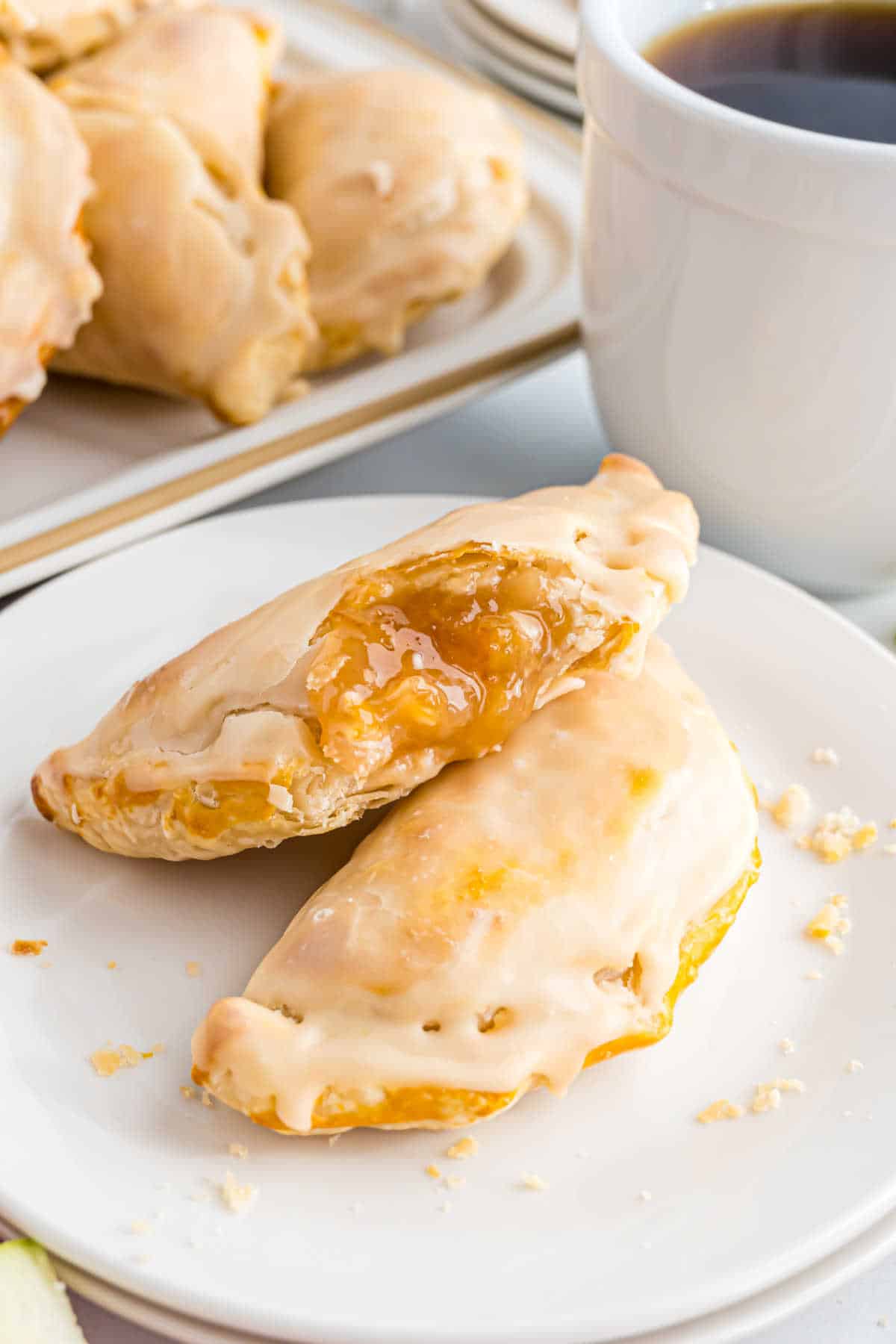 Two glazed apple empanadas with golden filling on a white plate, next to a cup of coffee.