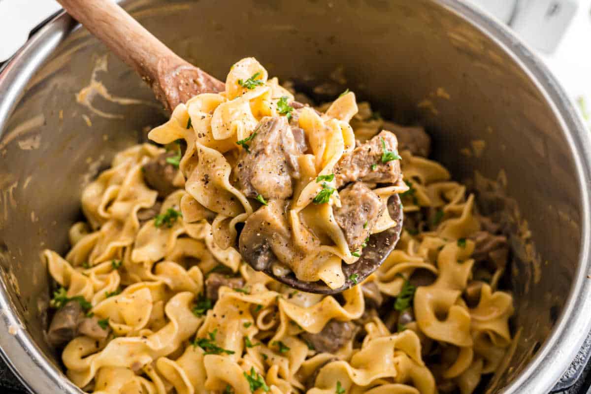 A wooden spoon lifts creamy beef stroganoff with egg noodles and parsley from a pot, showcasing the rich flavors of this classic dish.
