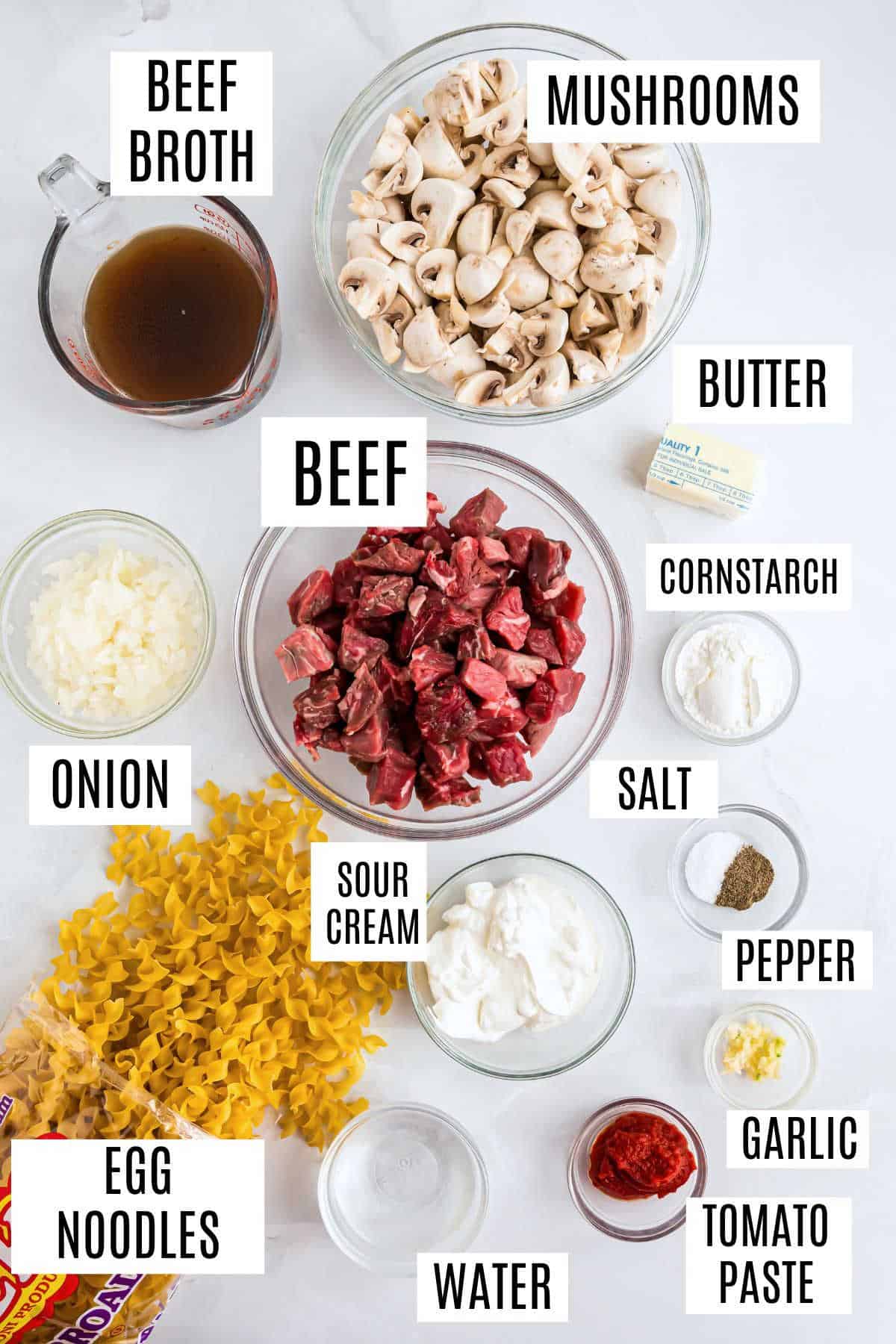 Ingredients for beef stroganoff laid out in bowls: tender beef, earthy mushrooms, noodles, savory broth, seasonings, and creamy dairy.