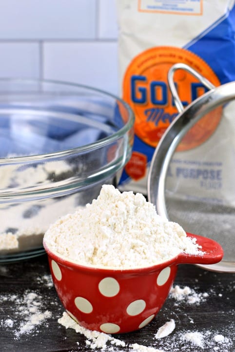 How do you measure flour properly? Do I need to sift it? Can I substitute unbleached flour for bleached flour? All your questions answered right here! Bag of flour with red and white polkadot measuring cup of flour.