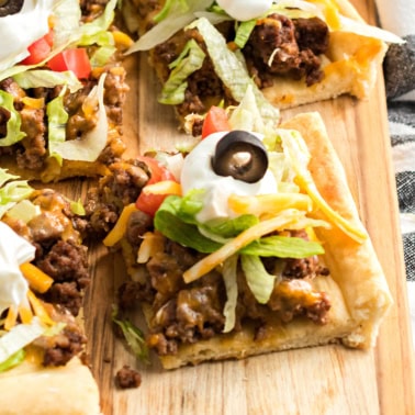 A flavorful taco pizza topped with seasoned ground beef, crisp lettuce, ripe tomatoes, melted cheese, tangy sour cream, and sliced olives sits enticingly on a wooden board.