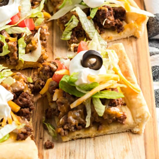 A flavorful taco pizza topped with seasoned ground beef, crisp lettuce, ripe tomatoes, melted cheese, tangy sour cream, and sliced olives sits enticingly on a wooden board.