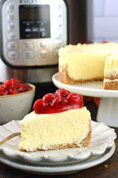 Instant Pot Cheesecake topped with cherry pie filling on white scalloped plate and instant pot in background.