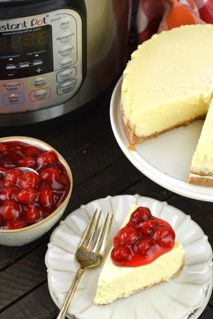 Slice of cheesecake with cherry filling. Whole cheesecake and instant pot in background.