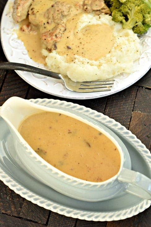 Gray gravy bowl filled with mushroom gravy with a white plate topped with mashed potatoes and pork chops in background.