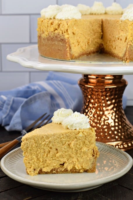 Pumpkin Cheesecake slice on white plate with whole cheesecake on cake plate.