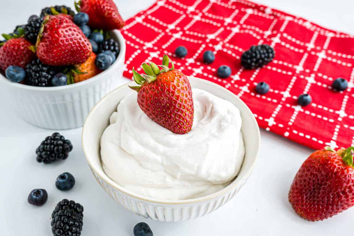 A bowl of homemade whipped cream topped with a strawberry, surrounded by berries and a red patterned cloth.