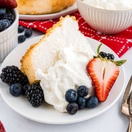 Slice of angel food cake topped with homemade whipped cream, blackberries, blueberries, and a sliced strawberry on a plate.