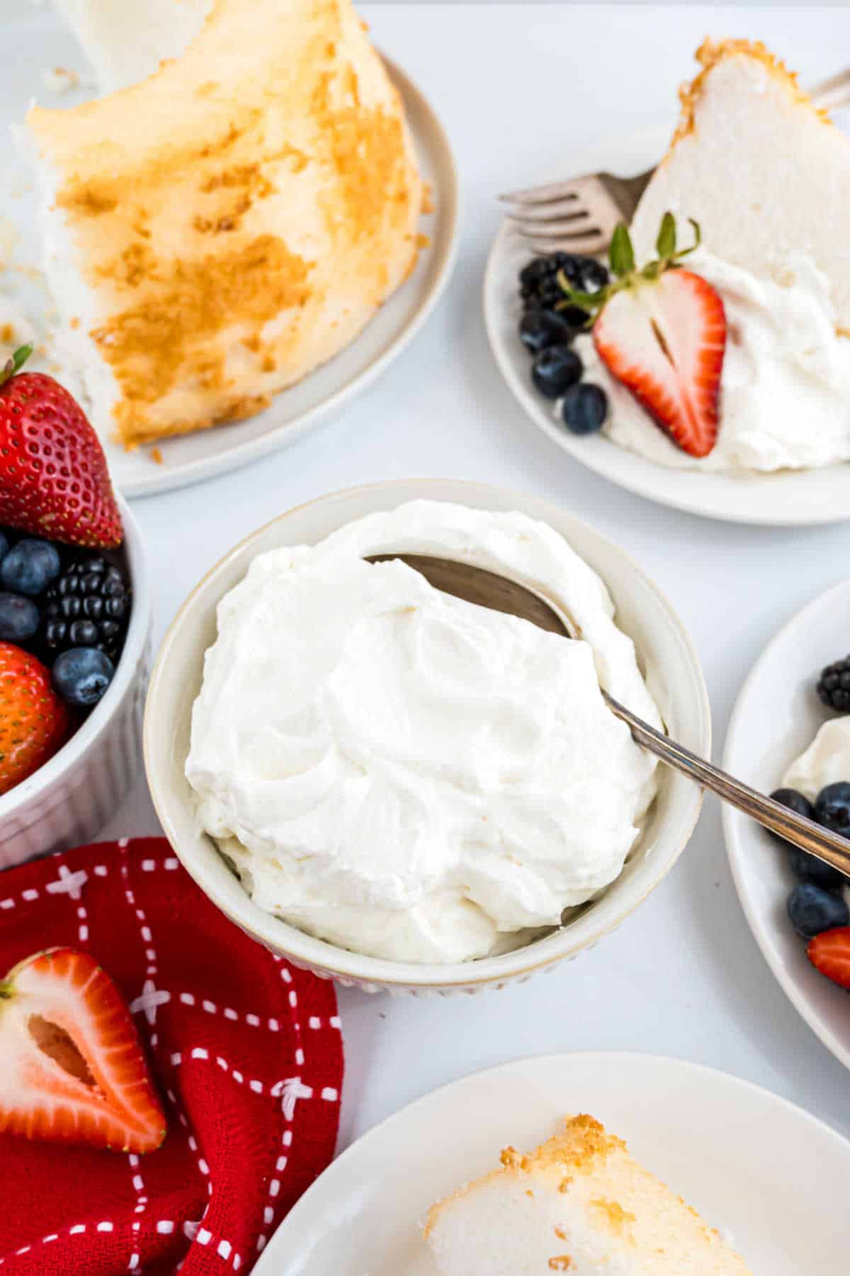 Bowl of homemade whipped cream with a spoon, surrounded by angel food cake slices and fresh berries on plates.