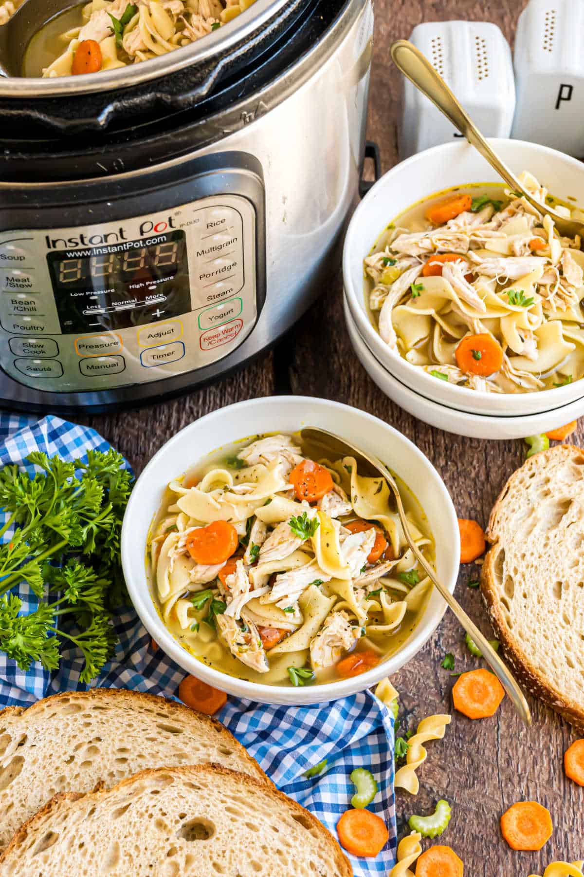Bowl of chicken noodle soup with carrots and bread sits beside an Instant Pot, garnished with parsley and accompanied by salt and pepper shakers.
