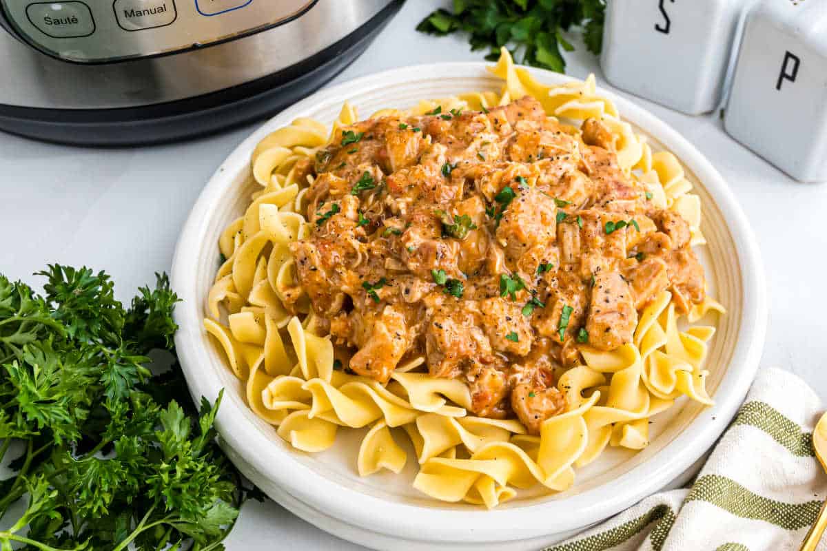A plate of egg noodles topped with creamy Instant Pot chicken paprikash, garnished with parsley, sits next to fresh herbs.