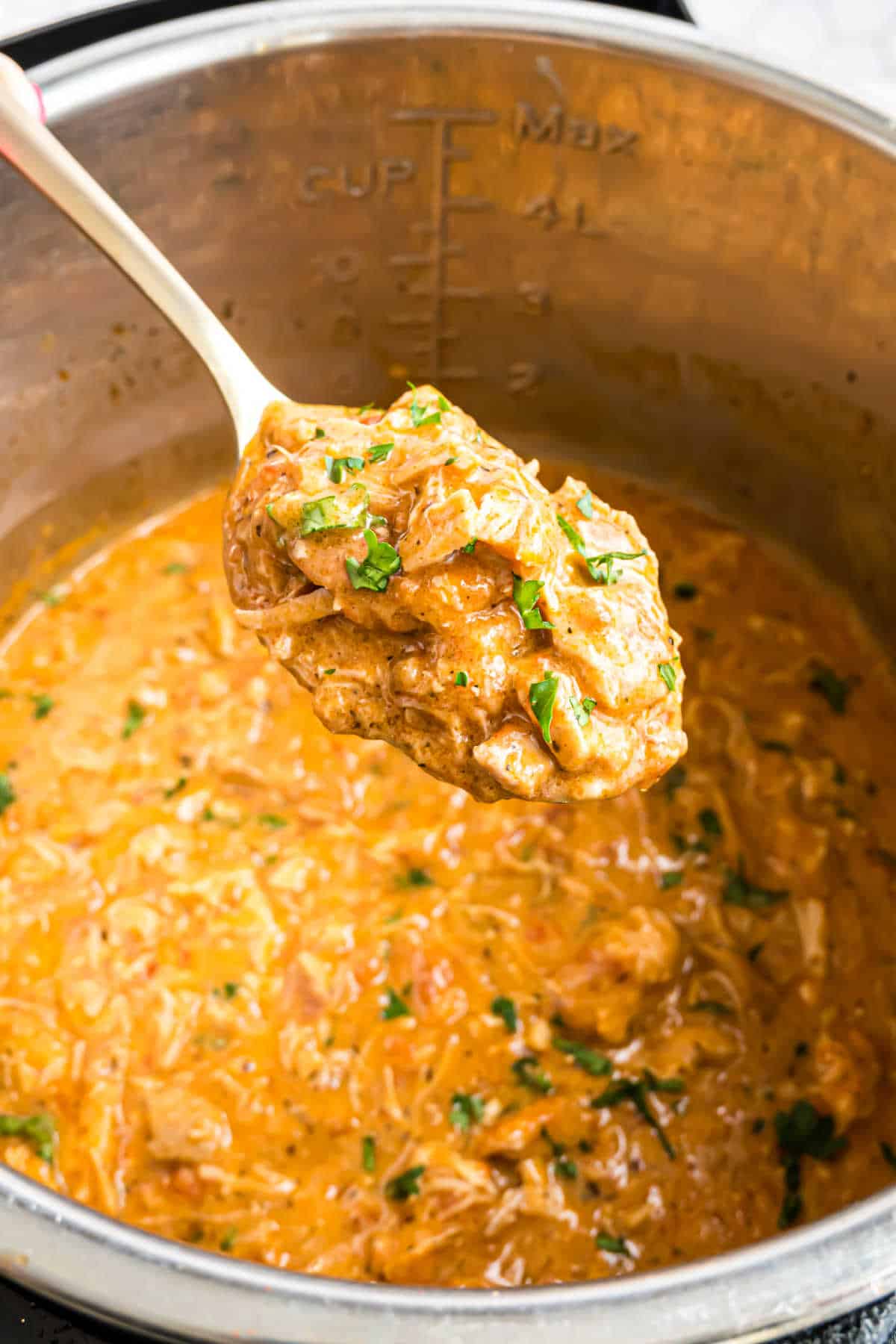 A spoon scoops creamy, shredded instant pot chicken paprikash in sauce from a metal pot, garnished with fresh herbs.