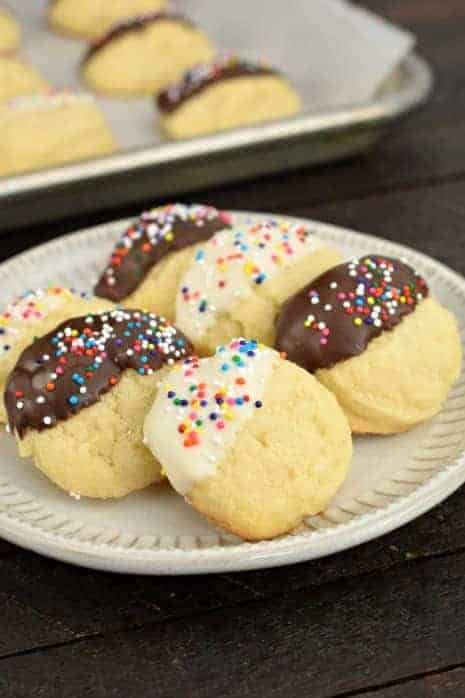 Plate of butter cookies dipping in white chocolate and some dipped in dark chocolate with sprinkles. Parchment paper pan of cookies in background.