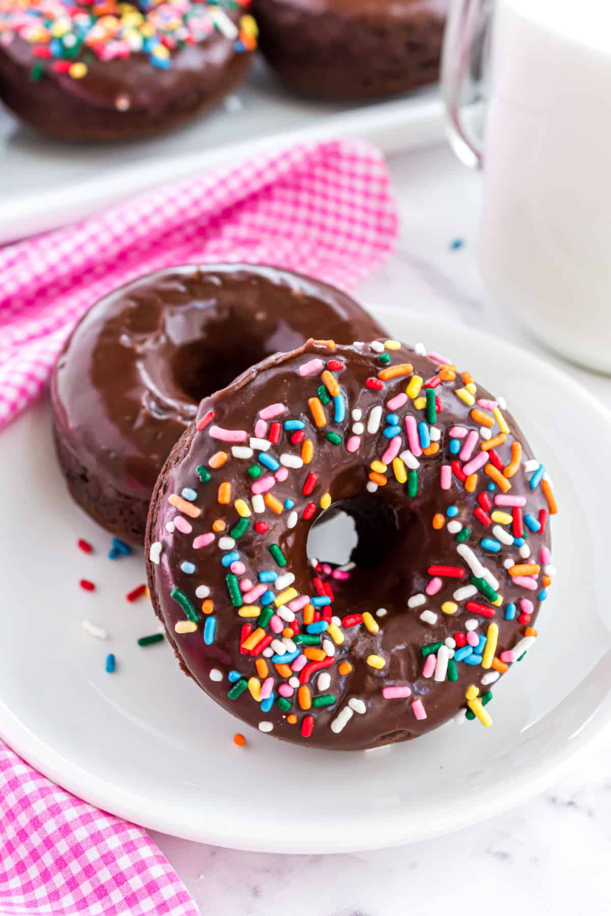 Two chocolate donuts on a white plate with pink gingham napkin.