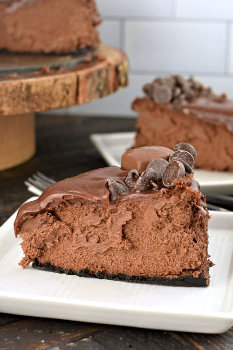 Rich, creamy, decadent Triple Dark Chocolate Cheesecake is the ultimate dessert. With my tips and tricks you can impress your friends and family with this chocolate cheesecake recipe! Big slice of dark chocolate cheesecake on white plate. Topped with chocolate ganache and chocolate chips