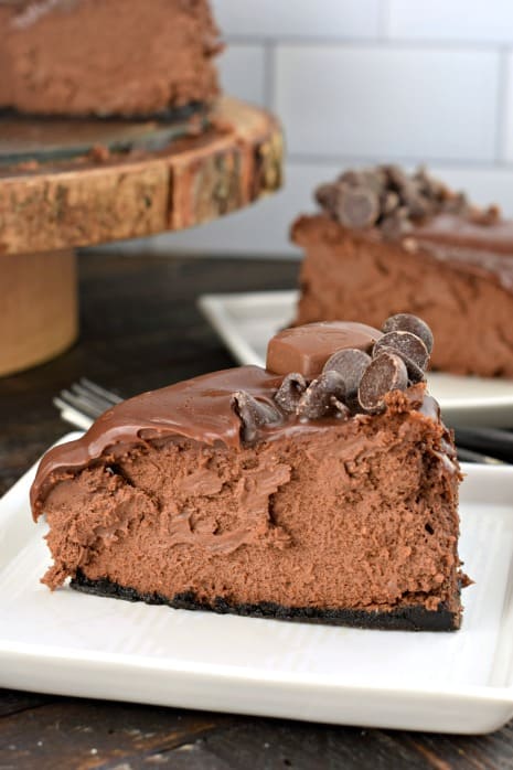 Big slice of dark chocolate cheesecake on white plate. Topped with chocolate ganache and chocolate chips