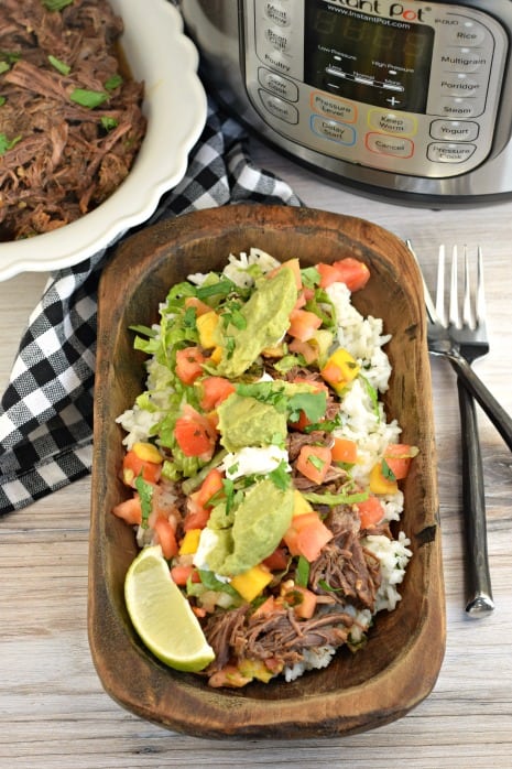 Wooden bowl with cilantro lime rice, beef barbacoa, guacamole, mango salsa, lime, and sour cream. Made in the instant pot.