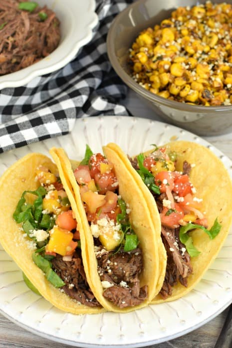 Three corn tortillas with instant pot beef barbacoa filling. Topped with mango salsa, lettuce, cotija cheese, and served with mexican corn.