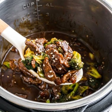A spoonful of instant pot beef broccoli in savory sauce, topped with sesame seeds, held over an Instant Pot.