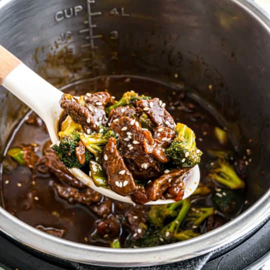 A spoonful of instant pot beef broccoli in savory sauce, topped with sesame seeds, held over an Instant Pot.