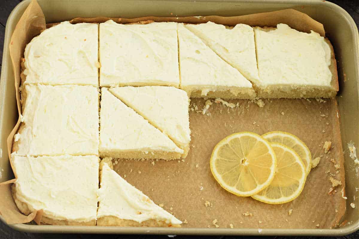 A tray of frosted lemon cake bars, partially sliced, with fresh lemon slices as garnish.