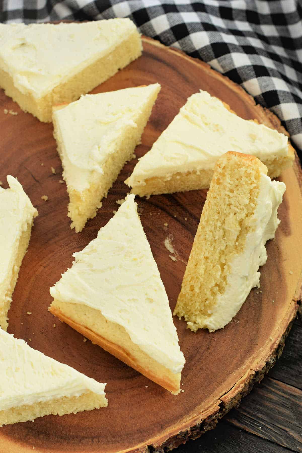 Triangular slices of frosted lemon cake bars rest invitingly on a wooden platter, with a checkered cloth adding a cozy backdrop.