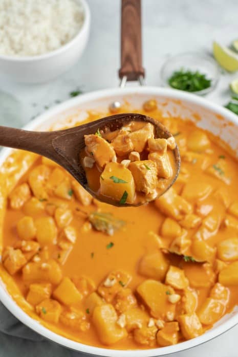 Wooden serving spoon lifting chicken curry out of white skillet.