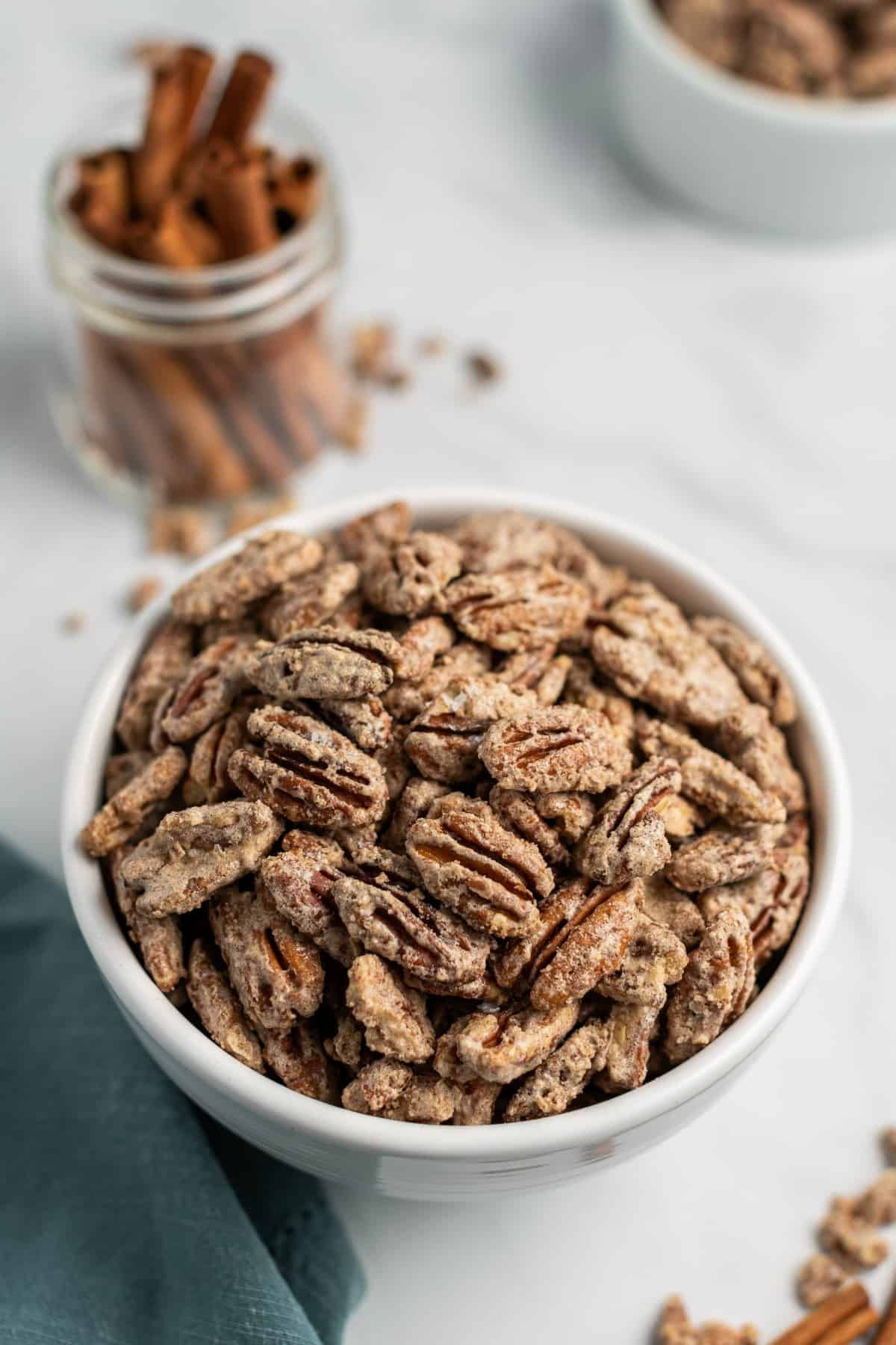 White bowl filled with candied pecans. Cinnamon sticks in background.