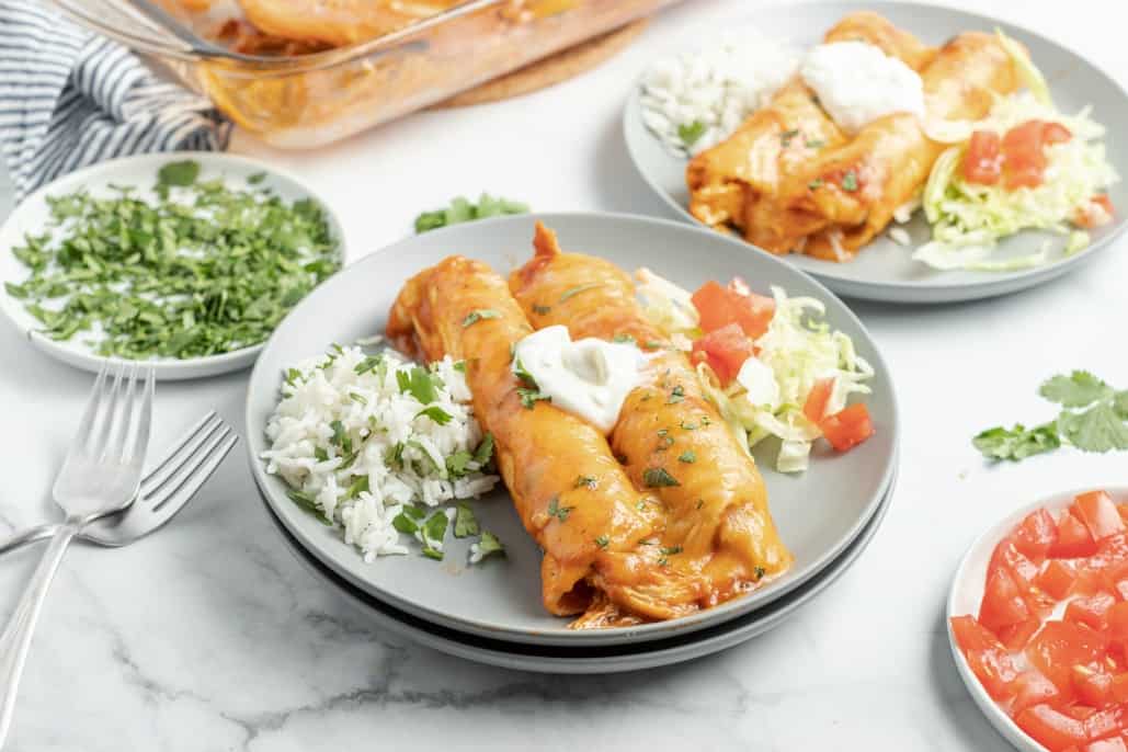 Two chicken enchiladas topped with sour cream and served with cilantro lime rice on a stack of gray plates.